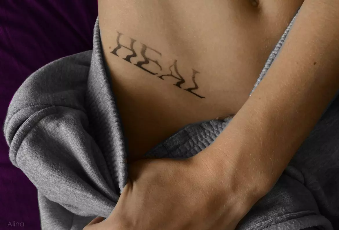 Bold black tribal-style lettering tattoo on the side rib, reading “Heal” in sharp, flame-inspired gothic script.